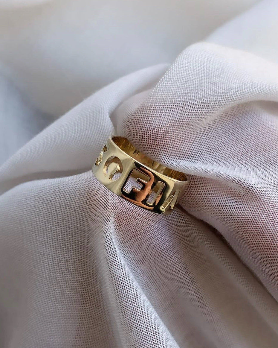 Letter Ring Wide in yellow gold with cut-out letters, resting on draped white fabric
