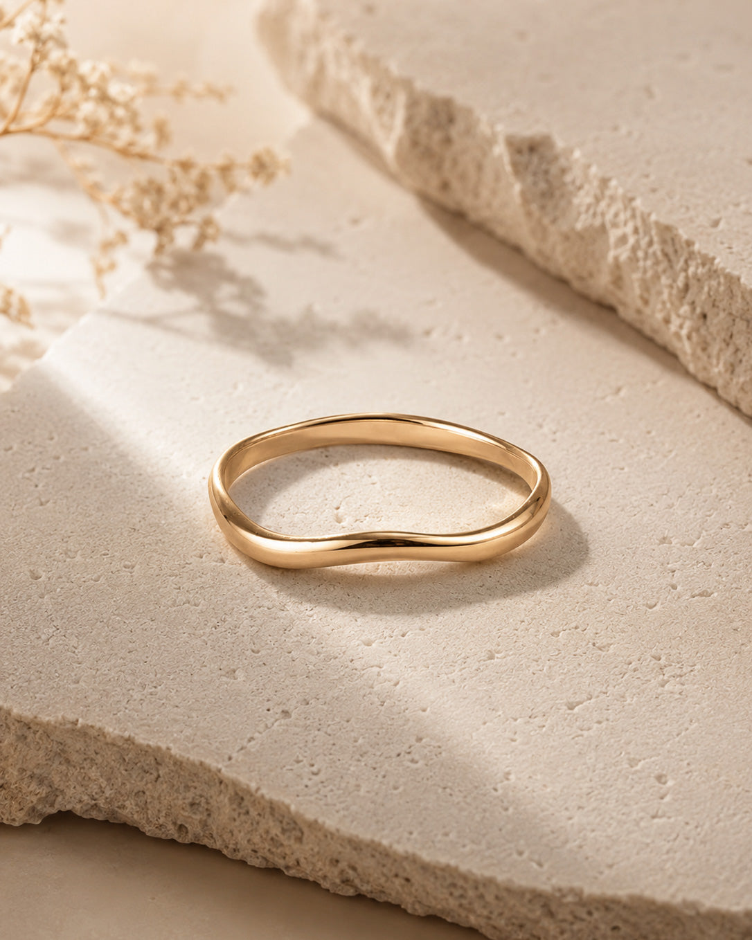 Lux Ring in yellow gold, a slim wavy band resting on a pale stone surface